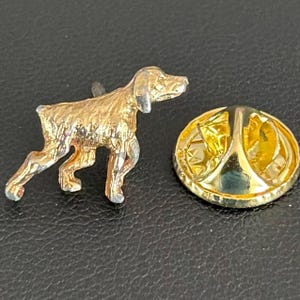 May include: A gold-colored dog pin with a silver-colored nose and paws. The pin is in the shape of a dog standing on all fours. The pin backing is gold-colored and has a pin clasp.