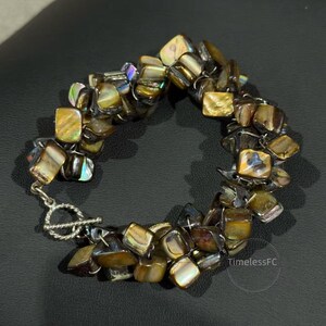 Op de afbeelding: Een handgemaakte armband met vierkante, iriserende kralen in goud, bruin en iriserende tinten. De armband is geregen met een zilverkleurige metalen ketting en sluiting. Het ontwerp is organisch en opvallend.