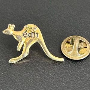 May include: A gold-colored metal pin in the shape of a kangaroo with the letters "edn" on its back. The pin has a clasp on the back. The pin is on a black surface.