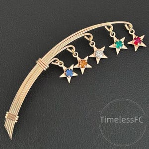 May include: Gold-toned brooch featuring a curved design with five dangling star-shaped charms. Each star holds a different colored gemstone: blue, orange, clear, green, and red. The brooch has a textured, detailed finish.