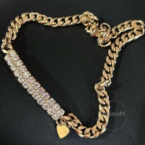 Op de afbeelding: Een goudkleurige ketting met een cubaanse schakelketting. De ketting heeft een gedeelte van heldere, sprankelende stenen en een kleine gouden hartvormige bedel. De sieraden worden op een zwart oppervlak weergegeven.