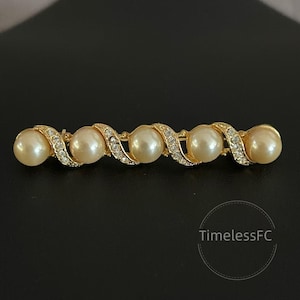 Può includere: Spilla dorata con cinque perle finte color crema. Le perle sono incastonate in un design ondulato con accenti di cristallo trasparente. La spilla è a barretta.