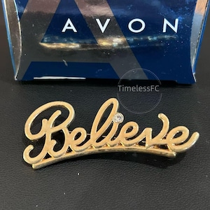 May include: A gold-coloured "Believe" brooch with a small, clear crystal. The word is in a cursive script. The brooch is set against a dark background, with a blue and white box in the background with the word "AVON".