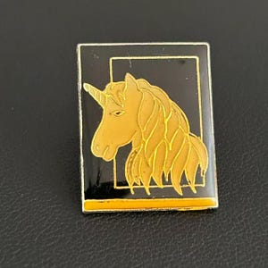 May include: A rectangular enamel pin featuring a gold-colored unicorn head profile set against a black background. The design is framed by a thin gold border, with a solid gold horizontal bar running across the bottom edge of the rectangular pin.