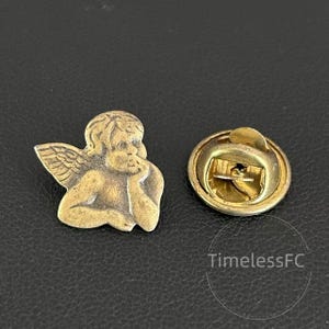 May include: A vintage-style lapel pin featuring a detailed cherub design in a bronze finish. The cherub is depicted with wings and is resting its chin on its hand. The pin has a classic butterfly clutch backing.