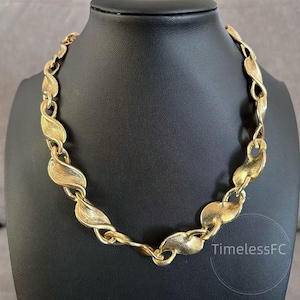 Puede incluir: Un collar dorado con un diseño en forma de hoja. El collar se exhibe en un maniquí negro. La cadena está hecha de eslabones interconectados, texturizados y curvados en forma de hoja, creando una pieza llamativa.