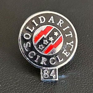 May include: A silver-toned pin with the words "SOLIDARITY CIRCLE" in a circular pattern around a central design. The central design features red and white stripes with three stars. The number "84" is at the bottom.