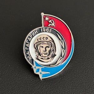 Puede incluir: Un pin metálico circular que presenta un retrato de Yuri Gagarin con casco espacial y el texto CCCP. El diseño incluye una sección roja con una hoz y un martillo, una sección azul con una estrella y el texto en cirílico YU.A. GAGARIN 1961.