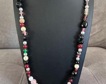 Puede incluir: Un collar de cuentas de colores con una variedad de formas y tamaños. El collar presenta cuentas en negro, rojo, rosa, blanco, plateado y verde. El collar se exhibe en un soporte negro.