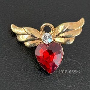 May include: A gold-toned angel wing pendant featuring a red heart-shaped gemstone and a clear crystal. The pendant has a loop for hanging and is set against a black backdrop. The text "TimelessFC" is visible.