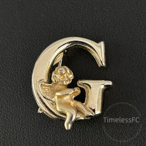 May include: Gold-coloured metal brooch in the shape of the letter "G" with a cherub figure. The cherub is seated within the curve of the letter. The brooch has a pin back. The text "TimelessFC" is in the lower right corner.