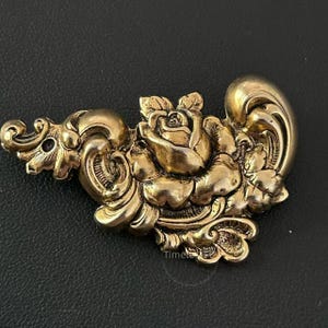 May include: Ornate gold-toned metal decorative piece featuring a central rose surrounded by detailed scrollwork and floral accents. The design has a vintage aesthetic, with a slightly tarnished finish, suggesting age or a distressed style. The piece is likely a decorative element.