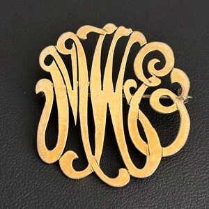 May include: A circular gold-toned brooch featuring an intricate, openwork monogram design with the intertwined letters M, W, and G. The metal surface has a smooth, polished finish with flowing, cursive-style lines forming the circular shape.