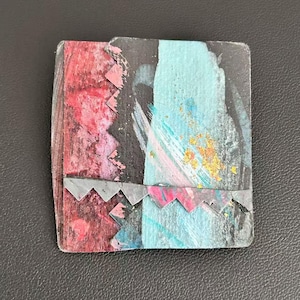 May include: A square brooch featuring a layered design with a red textured section on the left and a light blue painted area on the right. A dark gray zigzag metal trim separates the two sections, with gold-colored flecks on the blue surface.