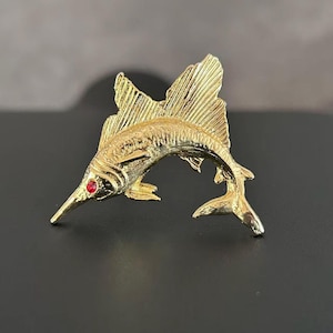 May include: A gold-toned metal brooch shaped like a leaping sailfish. The fish features a textured body, a prominent dorsal fin, and a single small, round, bright red gemstone set as the eye. The design captures the fish in a curved, dynamic pose.