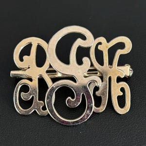 Könnte beinhalten: Goldfarbene Metallbrosche mit den Buchstaben "RGF" in Schreibschrift. Die Brosche hat einen Nadelverschluss auf der Rückseite und ist vor einem schwarzen Hintergrund platziert. Die Buchstaben sind verbunden und haben eine Vintage-Ästhetik.