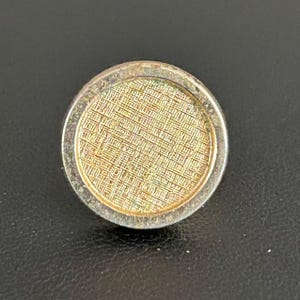 May include: A round, silver-toned button with a textured, gold-colored center. The center has a woven pattern. The button is likely for clothing or accessories.