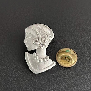 May include: A silver-toned metal brooch shaped like a stylized profile of a person with textured hair, a single clear rhinestone earring, and a beaded necklace. The back features a circular gold-toned metal pin clasp mechanism.