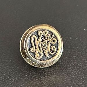 May include: A round, antique-style button with a gold-colored rim and a dark, ornate monogram in the center. The monogram appears to be the letters "MGC" in a stylized, swirling font. The button is set against a dark background.