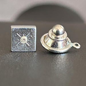 May include: Two silver-coloured metal accessories. One is a square cufflink with a central crystal and a starburst design. The other is a tie tack with a rounded top and a small loop. Both are on a dark surface.