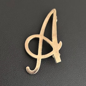 May include: A gold-toned metal brooch shaped like the capital letter A. The letter features a stylized, cursive-inspired font with a smooth, polished surface and a pointed top. The design includes an open loop on the left side and a curved tail.