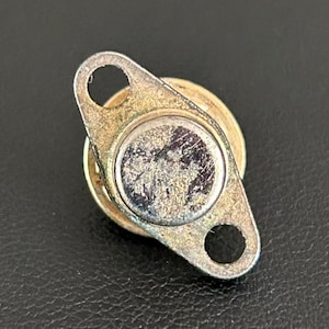 May include: A small, tarnished metal component with a circular base and a rectangular extension featuring two holes. The component has a silver-colored central disc and a gold-toned, aged appearance, suggesting it is vintage or used.