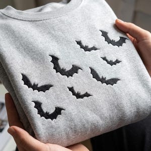 May include: A light gray crewneck sweatshirt featuring a pattern of small, black embroidered bats scattered across the front. The garment has a ribbed neckline and is made of a soft, heathered fabric with a consistent texture throughout the piece.
