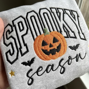 May include: A light grey sweatshirt featuring embroidered text that reads SPOOKY in large black block letters and season in black cursive script. An orange jack-o'-lantern with a green stem, two small black bats, and a gold star are embroidered.