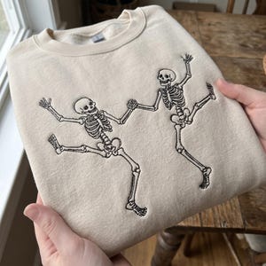 Könnte beinhalten: Ein beigefarbenes Sweatshirt mit Rundhalsausschnitt und einer schwarzen Stickerei von zwei tanzenden Skeletten. Die Skelette sind in einer dynamischen Pose mit erhobenen Armen und ausgestreckten Beinen dargestellt, während sie sich an den Händen halten.