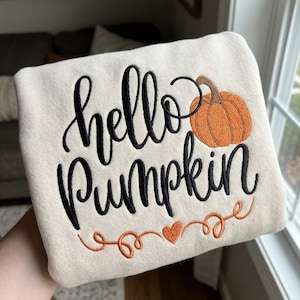 Könnte beinhalten: Ein cremefarbenes Sweatshirt mit schwarzer, gestickter Schreibschrift, die hello Pumpkin liest. Rechts neben dem Text befindet sich ein orangefarbener, gestickter Kürbis mit braunem Stiel. Unter dem Text ist ein orangefarbenes Herz mit Schnörkeln.