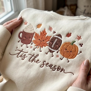 Könnte beinhalten: Ein cremefarbenes Sweatshirt mit bunter Stickerei, die einen lächelnden Kakaobecher, ein Ahornblatt, einen Football und einen Kürbis zeigt. Der Schriftzug 'tis the season' ist in brauner Schreibschrift unter den Motiven eingestickt.