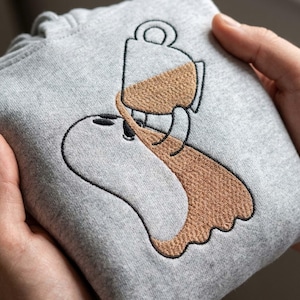 Könnte beinhalten: Ein hellgrauer Kapuzenpullover mit einer Stickerei, die einen weißen Geist zeigt, der sich eine Tasse braunen Kaffee über sich selbst gießt. Die Stickerei verwendet schwarze Umrisse für den Geist und die Tasse sowie braunes Garn.