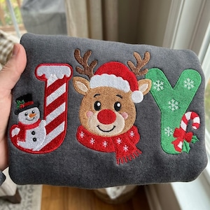 May include: A dark gray sweatshirt featuring embroidered holiday designs spelling JOY. The letter J is a red and white candy cane with a snowman, the O is a reindeer wearing a Santa hat and red scarf, and the Y is green with snowflakes and a candy cane.