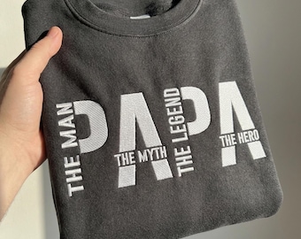 May include: A charcoal gray crewneck sweatshirt featuring large white embroidered text that reads PAPA. Smaller white text integrated into the design reads THE MAN, THE MYTH, THE LEGEND, and THE HERO, arranged vertically and horizontally.
