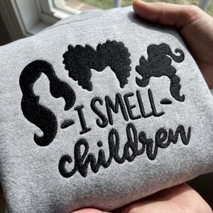 Puede incluir: Una sudadera gris claro con bordado negro de tres siluetas de cabello distintas dispuestas sobre el texto I SMELL children. El bordado está centrado en la tela, mostrando costuras detalladas tanto para las formas del cabello como para las letras.