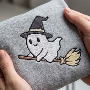 Puede incluir: Una tela gris jaspeado con un diseño bordado de un pequeño fantasma blanco que lleva un sombrero de bruja negro con una banda beige. El fantasma aparece volando sobre una escoba de madera marrón con cerdas beige, en un estilo sencillo.