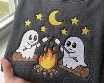 May include: A charcoal gray hooded sweatshirt featuring an embroidered design of two white ghosts sitting on brown logs by a campfire. The ghosts are roasting white marshmallows on sticks under a yellow crescent moon and five yellow stars.