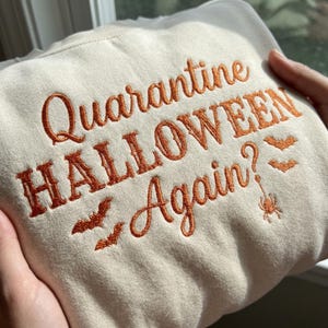 May include: A cream-colored sweatshirt featuring orange embroidered text that reads "Quarantine HALLOWEEN Again?" The design includes small embroidered orange bats and a spider hanging from a web, all stitched onto the front of the garment.