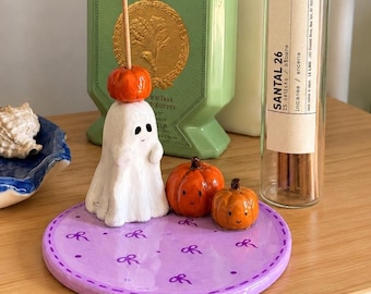 May include: A round lavender ceramic tray decorated with small purple bows and dots, featuring a white ghost figurine with a small orange pumpkin on its head, and two additional orange pumpkins with painted faces sitting on the tray surface.