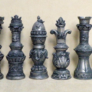 Puede incluir: Un conjunto de seis piezas de ajedrez ornamentadas de color plateado. Cada pieza tiene un diseño único, que incluye coronas, flor de lis y tallas decorativas. Las piezas están dispuestas en fila, mostrando su detallada artesanía.