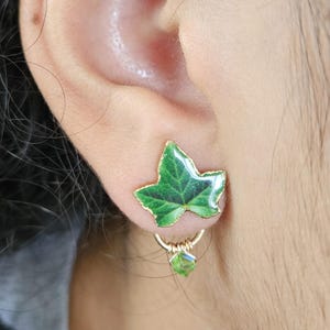 May include: A gold-coloured earring with a green leaf design and a gold outline. The leaf has detailed veining and a small, dangling green crystal. The earring is attached to the ear with a gold hoop.