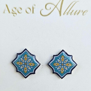 Puede incluir: Un par de pendientes cuadrados con un diseño floral turquesa y amarillo. Los pendientes tienen un borde azul oscuro y están sobre un fondo blanco. El texto "Age of Allure" está en la parte superior.