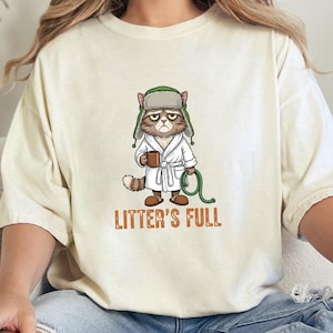 May include: Cream-colored t-shirt featuring a grumpy cat illustration. The cat is wearing a bathrobe and a hat, holding a mug and a green hose. The text "LITTER'S FULL" is printed below the cat.