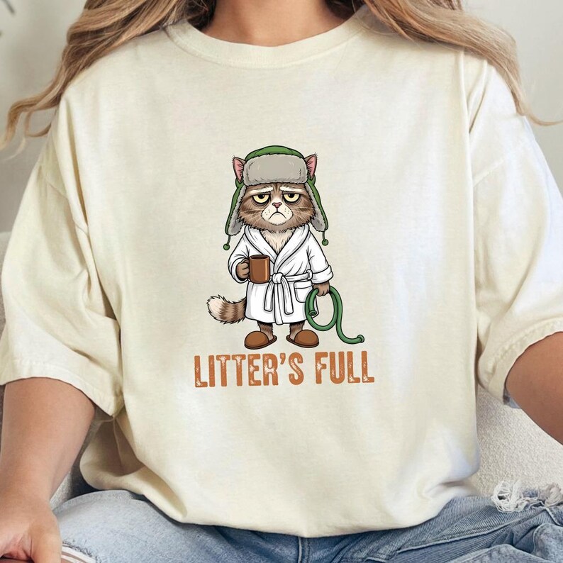 May include: Cream-colored t-shirt featuring a grumpy cat illustration. The cat is wearing a bathrobe and a hat, holding a mug and a green hose. The text "LITTER'S FULL" is printed below the cat.