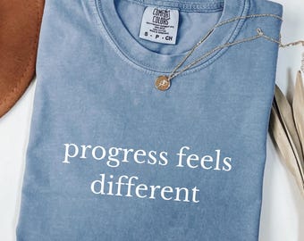 Może przedstawiać: Niebieska koszulka Comfort Colors z białym napisem "progress feels different". Złoty naszyjnik z wisiorkiem leży na złożonej koszulce. Rozmiar S.