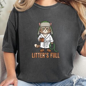 May include: Dark gray t-shirt featuring a grumpy cat illustration wearing a bathrobe and hat, holding a mug and a green hose. The text "LITTER'S FULL" is printed in orange below the cat.