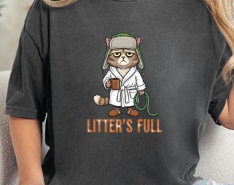 May include: Dark gray t-shirt featuring a grumpy cat illustration wearing a bathrobe and hat, holding a mug and a green hose. The text "LITTER'S FULL" is printed in orange below the cat.