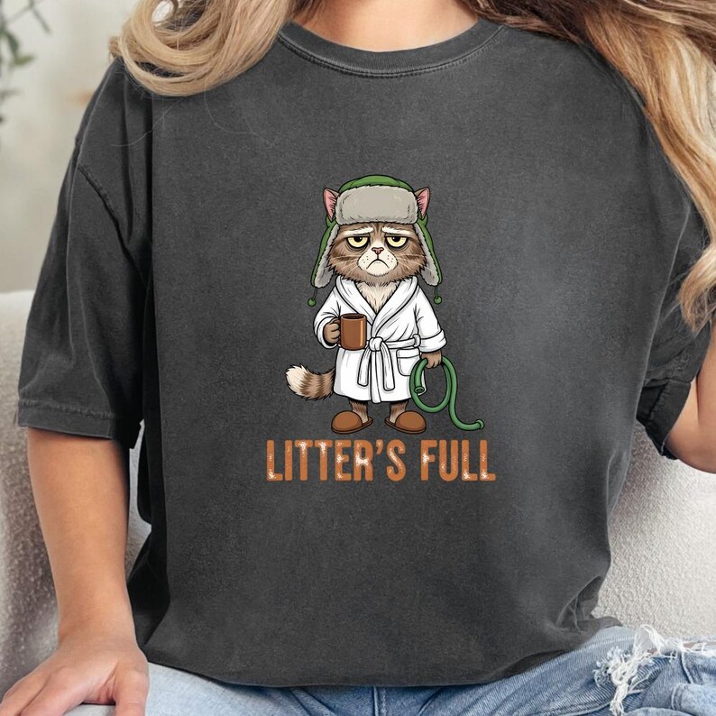 May include: Dark gray t-shirt featuring a grumpy cat illustration wearing a bathrobe and hat, holding a mug and a green hose. The text "LITTER'S FULL" is printed in orange below the cat.