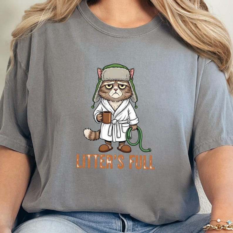 May include: Gray t-shirt featuring a grumpy cat wearing a bathrobe and a hat, holding a mug and a green hose. The text "LITTER'S FULL" is printed in orange below the cat illustration.