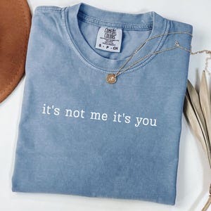 Puede incluir: Una camiseta azul Comfort Colors con la frase "it's not me it's you" en blanco. Un collar dorado con colgante está sobre la camiseta doblada. La camiseta está sobre una superficie blanca.